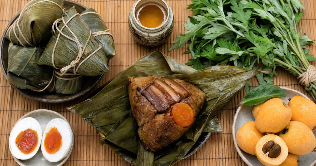 Bamboo mat with pyramid sticky-rice dumplings wrapped in green leaves and twine, one unwrapped showing rice with pork and yolk, salted duck eggs, a jar of wine, mugwort.