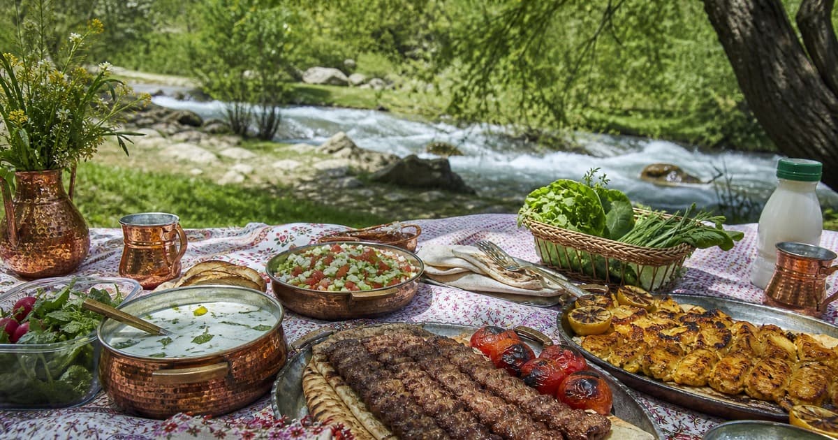 Copper pots, yogurt soup, salad, grilled kebabs, tomatoes, potato frittata, herb basket, radishes and milk bottle on a cloth by a river