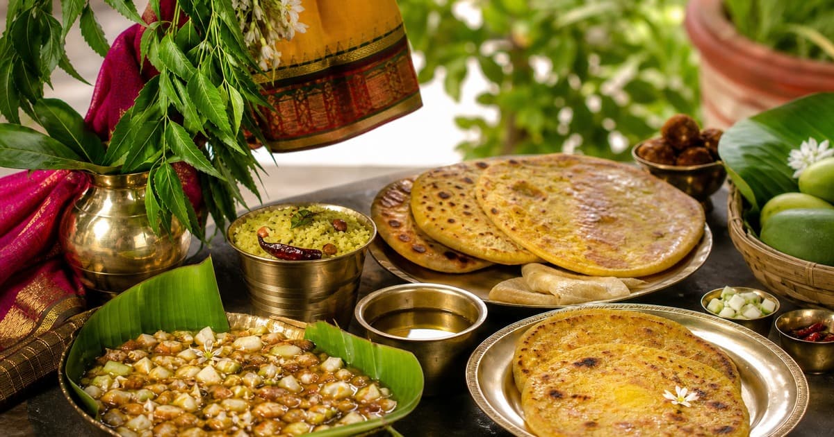 Banana-leaf bowl of mixed beans, stacked flatbreads, brass bowls, a pot with green leaves, mangoes in a basket, and small sweets on a table